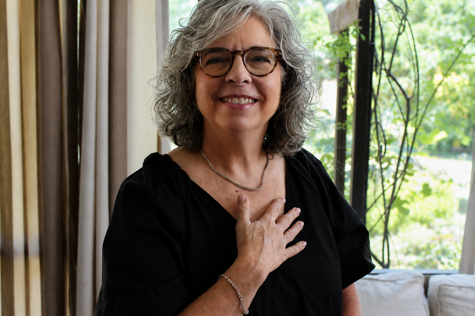 Smiling woman with glasses wearing a silver necklace and bracelet, hand resting on her chest